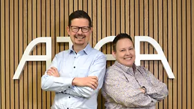 Jarno Oksanen ja Petra Berg seisovat vierekkäin ja hymyilevät. Taustalla puisella seinällä on valkoinen Atean logo.