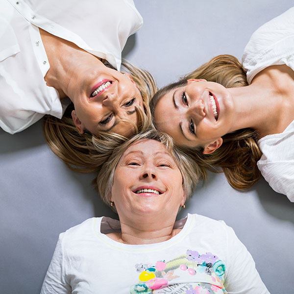 Kolme eri sukupolviin kuuluvaa naista makaa selällään niin, että heidän päälakensa koskettavat.
