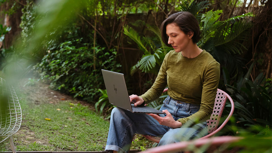 Nainen istuu ulkona puutarhassa ja työskentelee hp-merkkisellä kannettavalla tietokoneella.