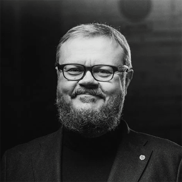 Hymyilevä pikkutakkiin ja mustaan poolopaitaan pukeutunut mies katsoo kameraan, tausta on tumma ja neutraali.