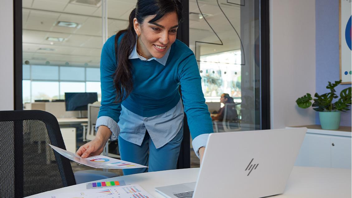 Nainen seisoo pöydän ääressä kannettavan tietokoneen ääressä paperi kädessä.