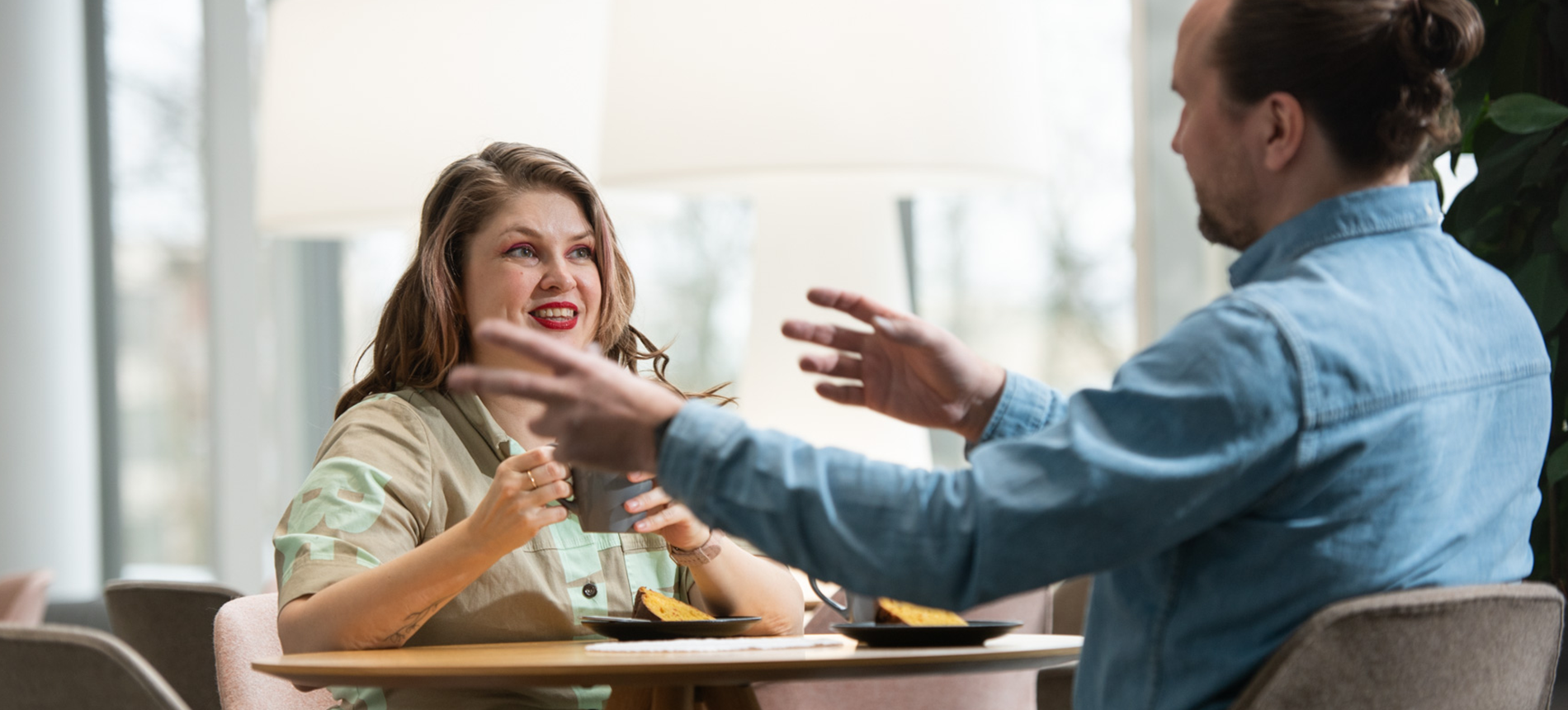 Myrsky Tammimaa ja Tomi Tihekari istuvat kahvilapöydässä. Tomi elehtii käsillään ja Myrsky kuuntelee.