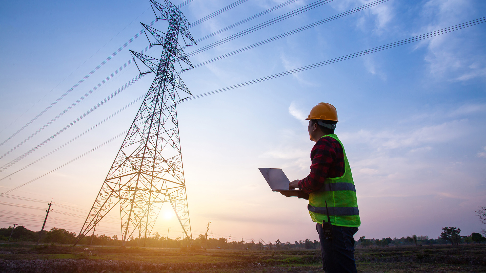 Huomioliiviin ja työmaakypärään pukeutunut henkilö seisoo kannettava tietokone kädessään ja katsoo voimajohtopylvästä.