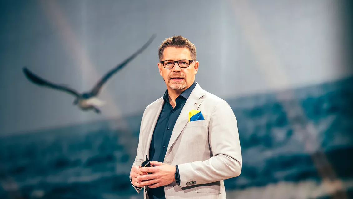 Juha Sihvonen seisoo Atea Focus -tapahtuman päälavalla puhumassa. Taustalla on kuva lentävästä lokista.
