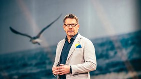 Juha Sihvonen seisoo Atea Focus -tapahtuman päälavalla puhumassa. Taustalla on kuva lentävästä lokista.