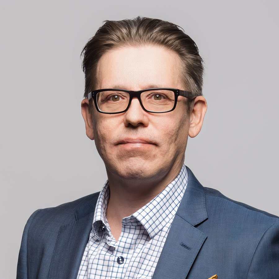 Mies, joka on pukeutunut keskisiniseen puvuntakkiin ja ruudulliseen kauluspaitaan ja jolla on keskiruskeat, taaksepäin kammatut hiukset ja silmälasit.